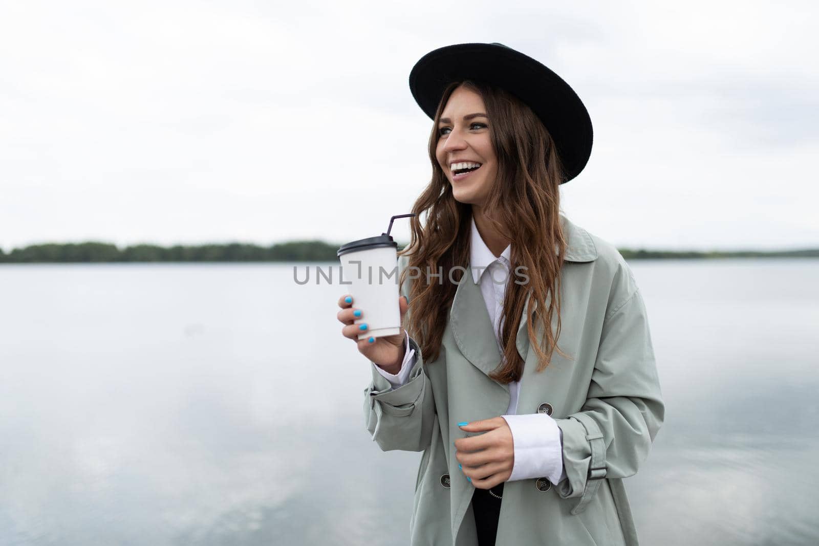 a young woman walks along the embankment against the backdrop of a large lake with a glass of coffee with a smile and in a black hat.