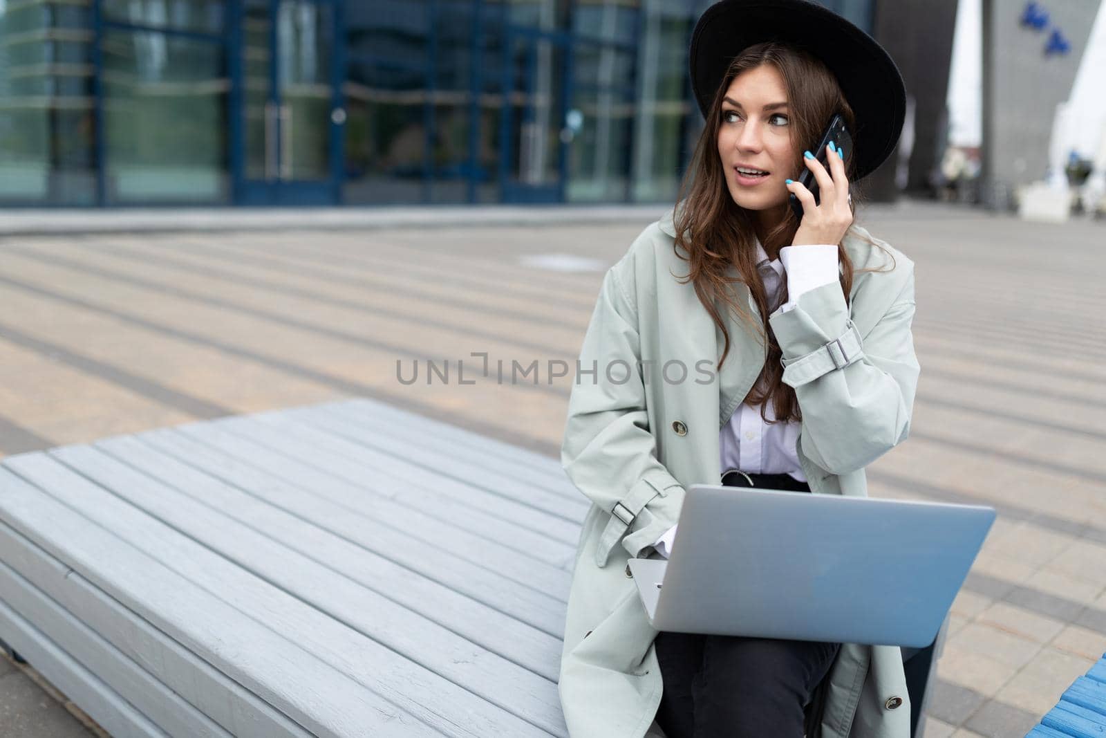 young woman works online on a laptop and speaks on a mobile phone Sitting on a bench next to an office building.