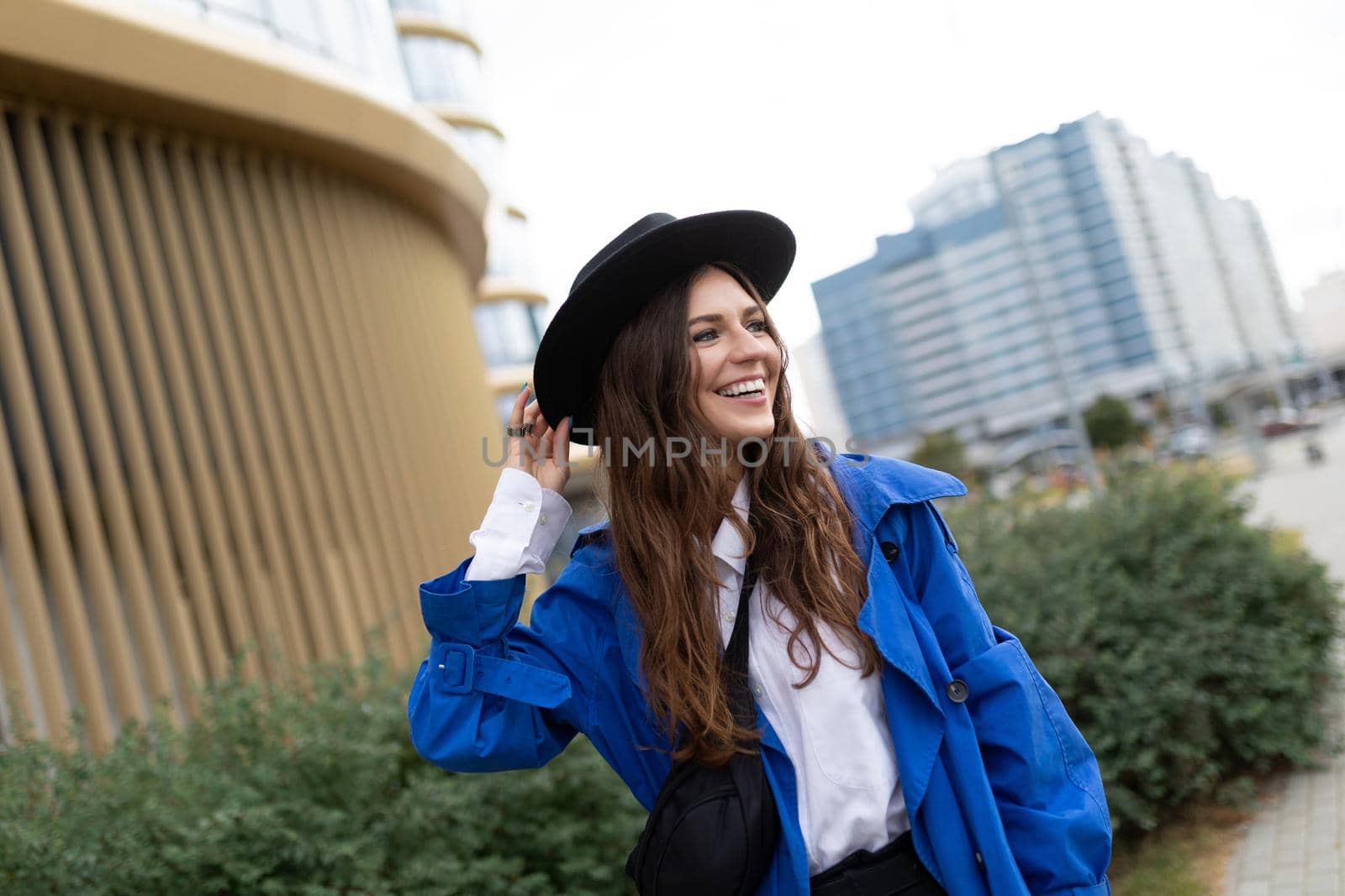 portrait of a strong young woman in a black hat and blue raincoat walking around the city with a smile on her face.