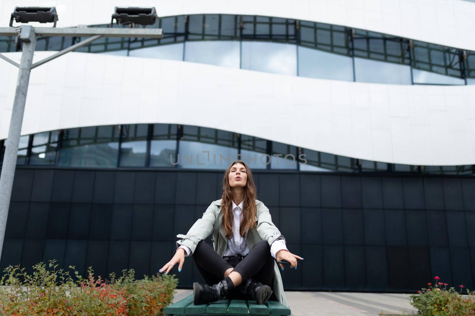 a young woman sits in a yoga position against the backdrop of an office building on a bench with her eyes closed, arms spread apart.