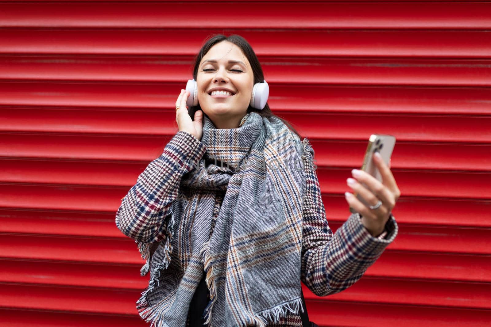 young and happy woman in headphones closing her eyes listens to music with a wide smile on her face through a mobile phone against the background of a red bright wall.