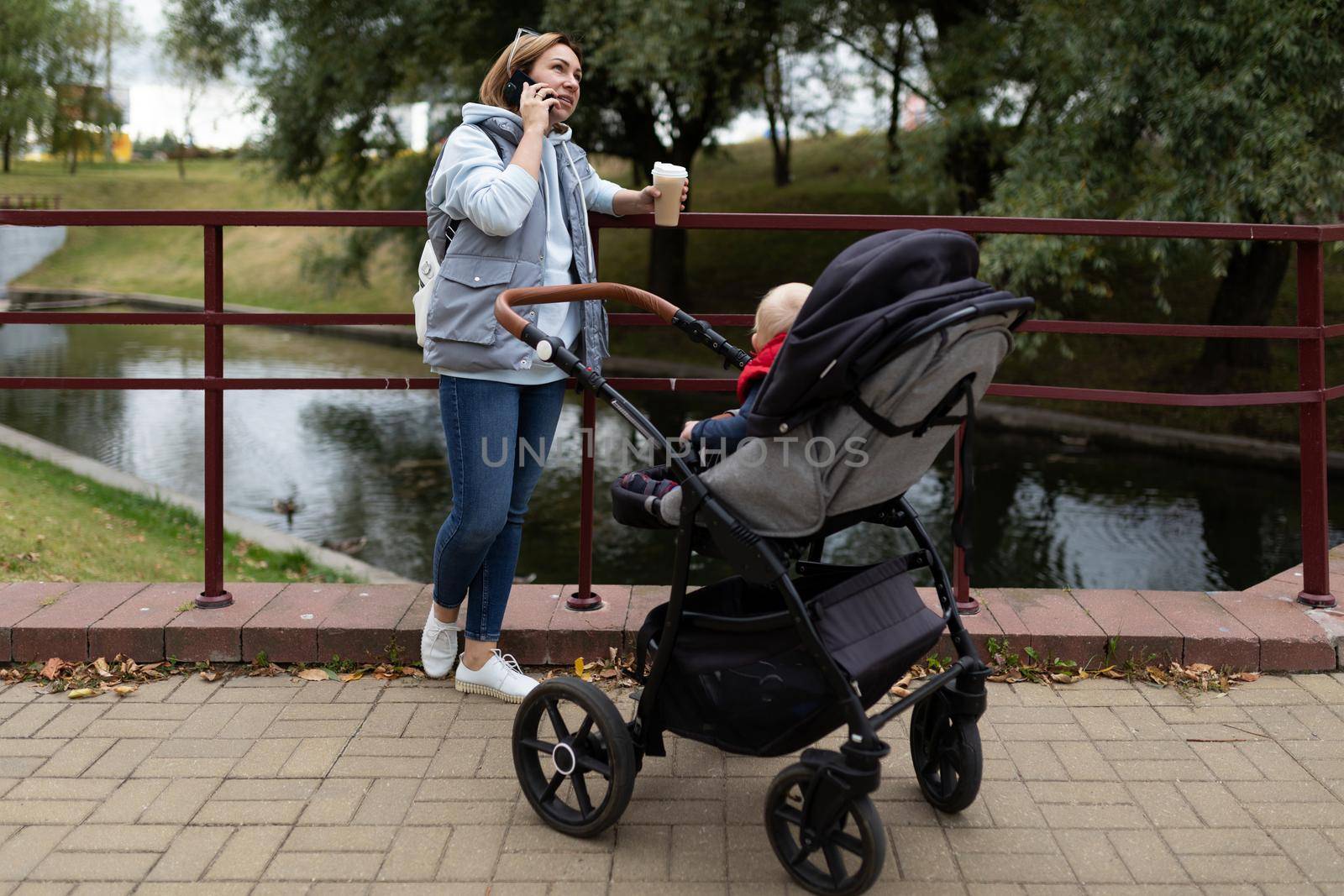 young caring mother with a cup of coffee in her hands next to the stroller with her newborn child while walking in the park speaks on the phone with a smile on her face.