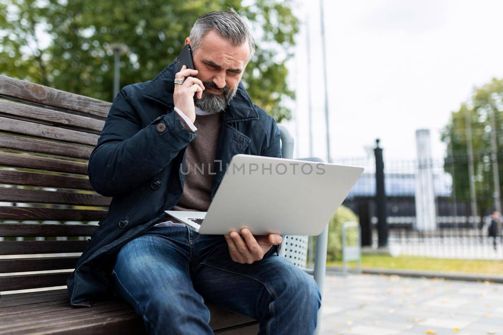 on the way to the office, a man is talking on the phone and looking at a laptop while sitting on a bench next to the business center.