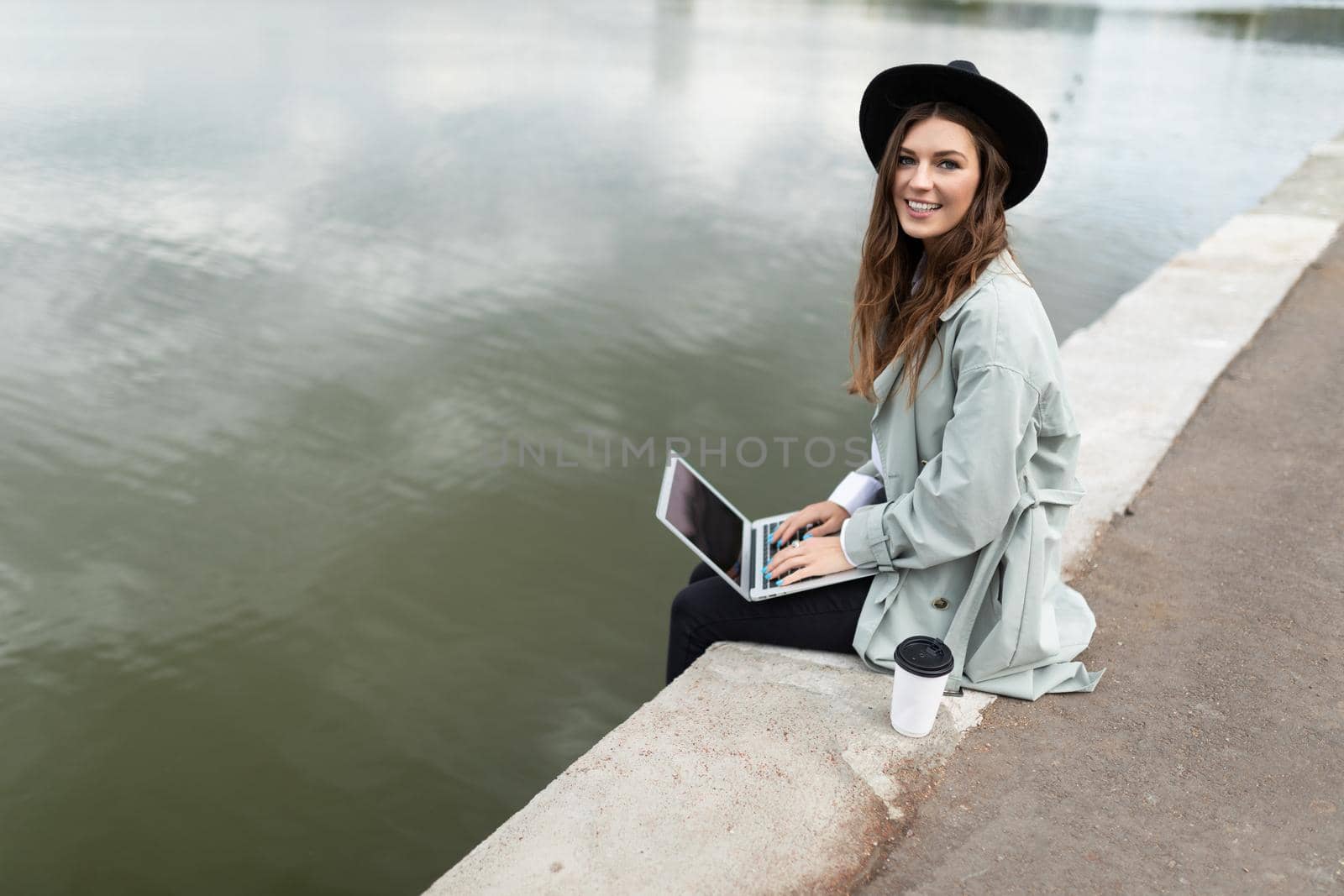 young woman working on a laptop online sitting on the promenade dangling her legs to the water with a smile looking at the camera.