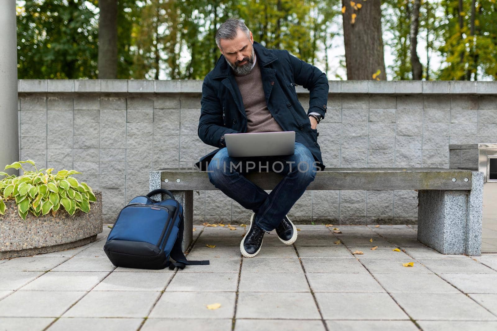 mature businessman man working online on a laptop sitting on a bench in a city park.