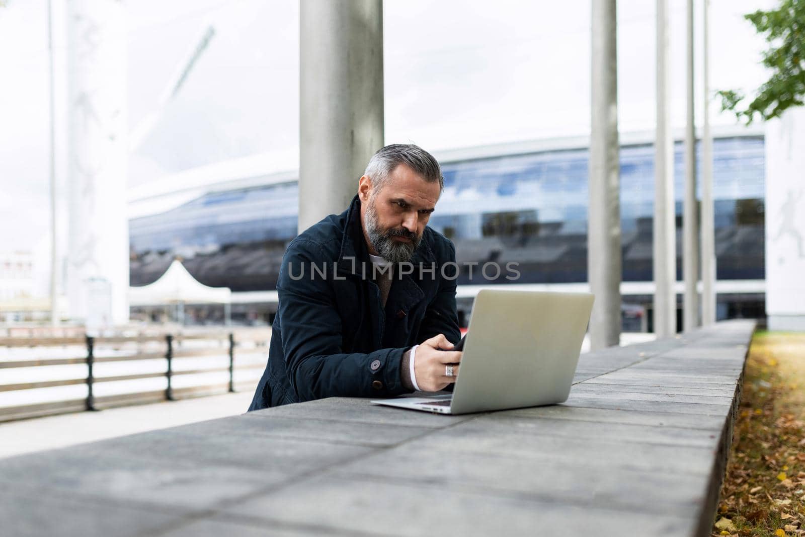 businessman man with a gray beard works on a laptop in a city park against the backdrop of an office building.