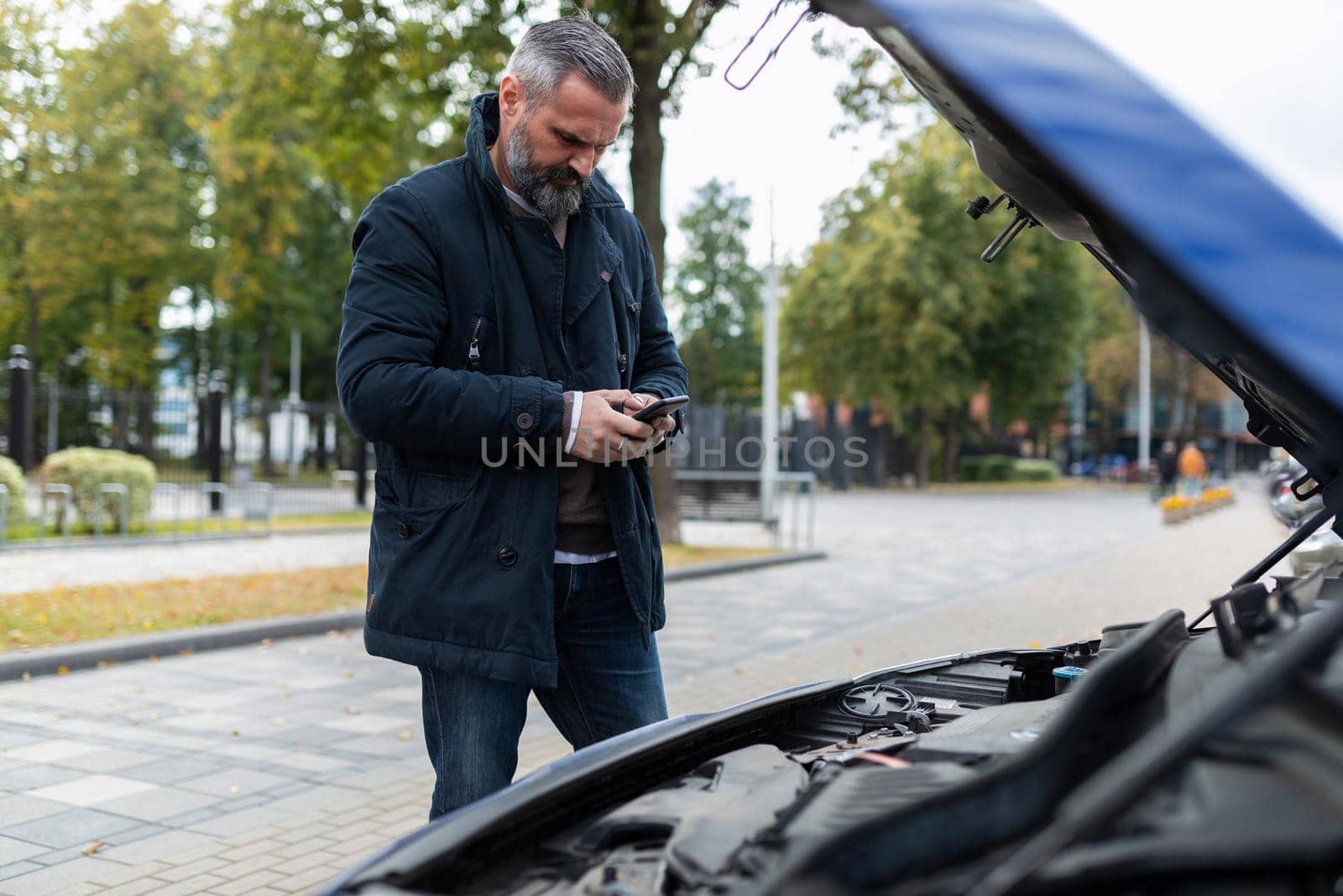an elderly man next to the raised hood of a broken car calls for technical support on a mobile phone.