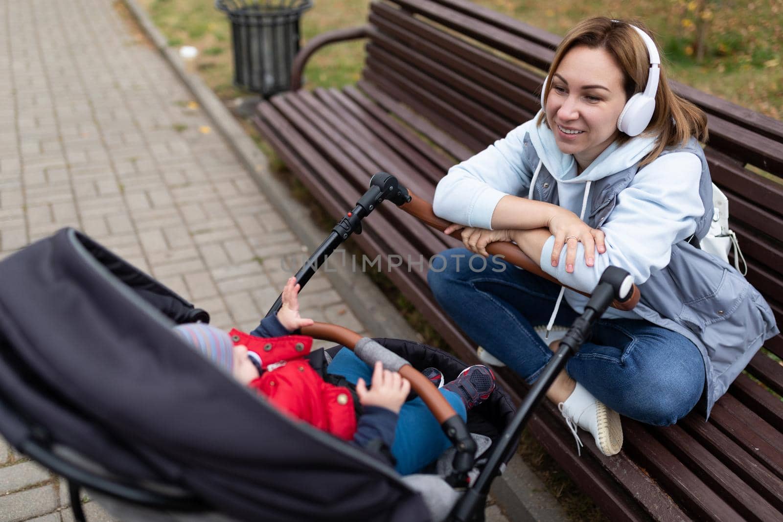 young woman sitting on a bench listening to music with headphones next to a pram with her baby.
