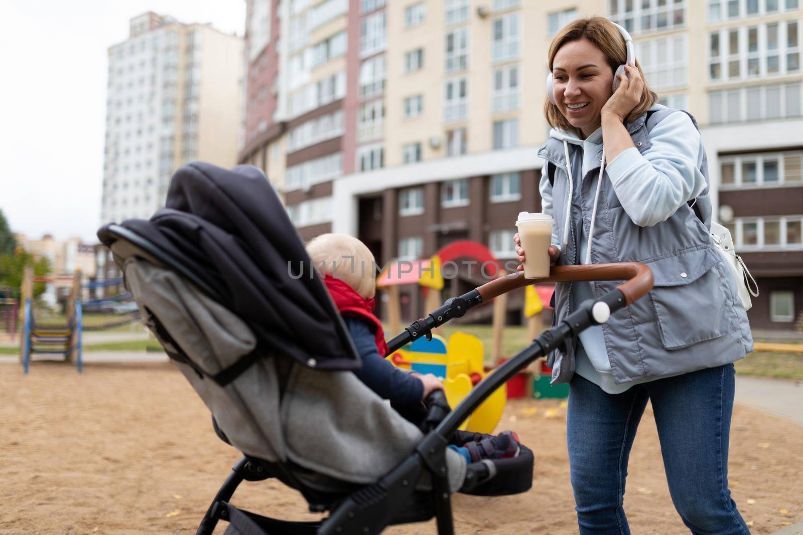 a young mother listens to music on headphones and drinks coffee while walking with a baby in a pram on a playground near the house by TRMK