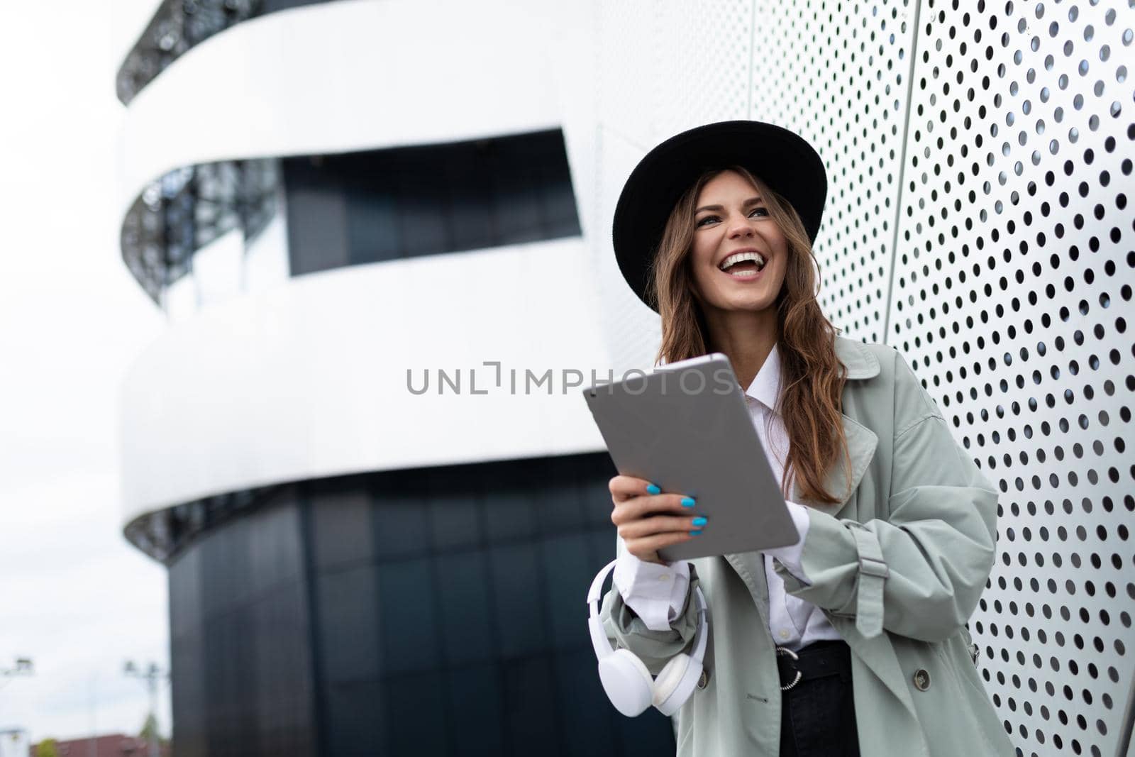 portrait of a young woman in a hat and an autumn gray coat against the background of an aluminum facade of an office building with a tablet in her hands and a smile on her face.