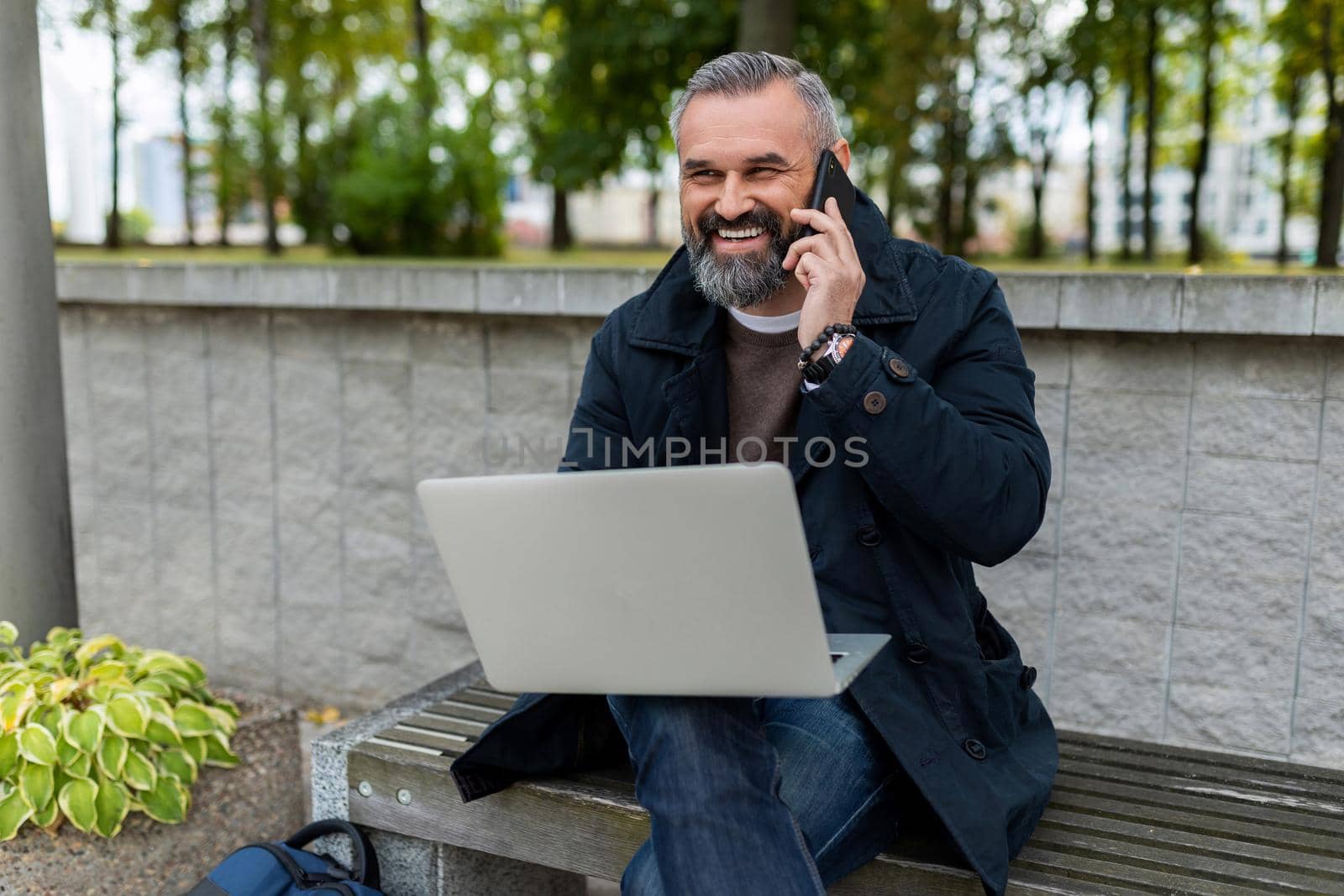 mature male entrepreneur working online on a laptop in a park in a city talking on the phone with a smile.