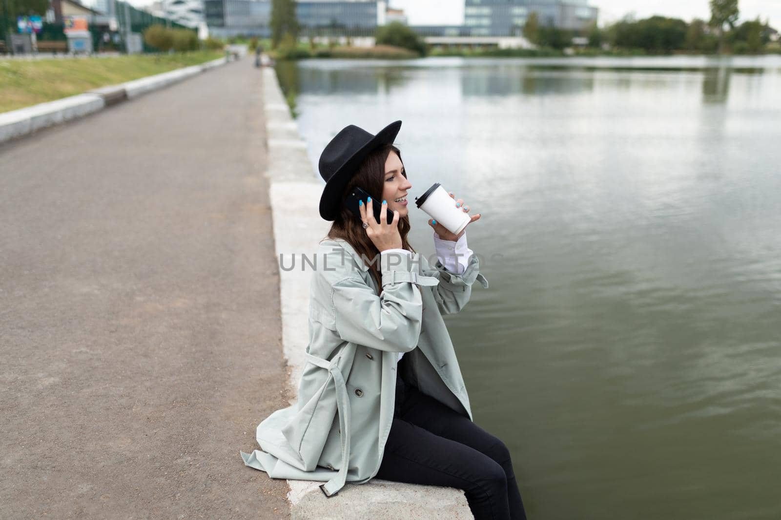 a young woman sits on the embankment dangling her legs to the water and drinking coffee talking on the phone looks into the distance.