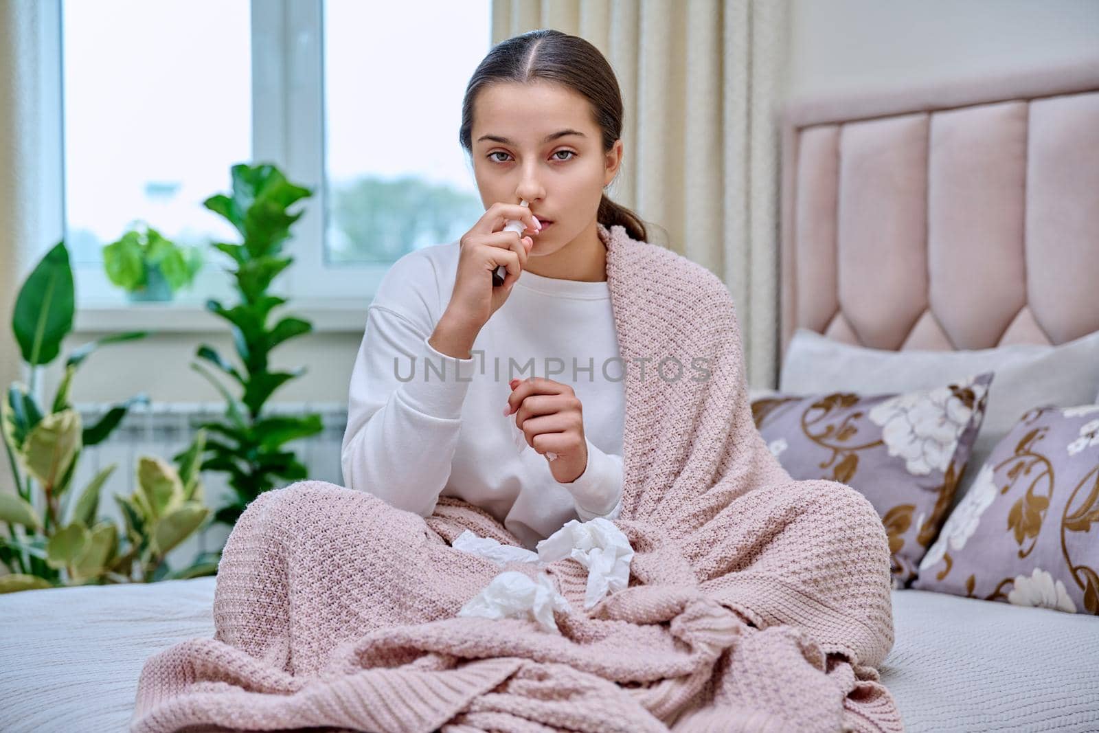 Young teenage female sitting in bed with runny nose medicine spray drops, sneezing into tissues, burying her nose. Autumn winter, rhinitis runny nose stuffy nose, treatment, flu season