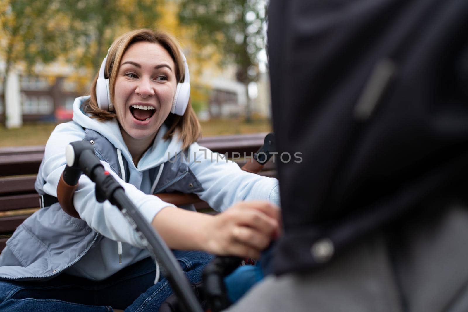 a young mother outdoors sitting on a bench listens to music on headphones and emotionally plays with her baby in a stroller by TRMK