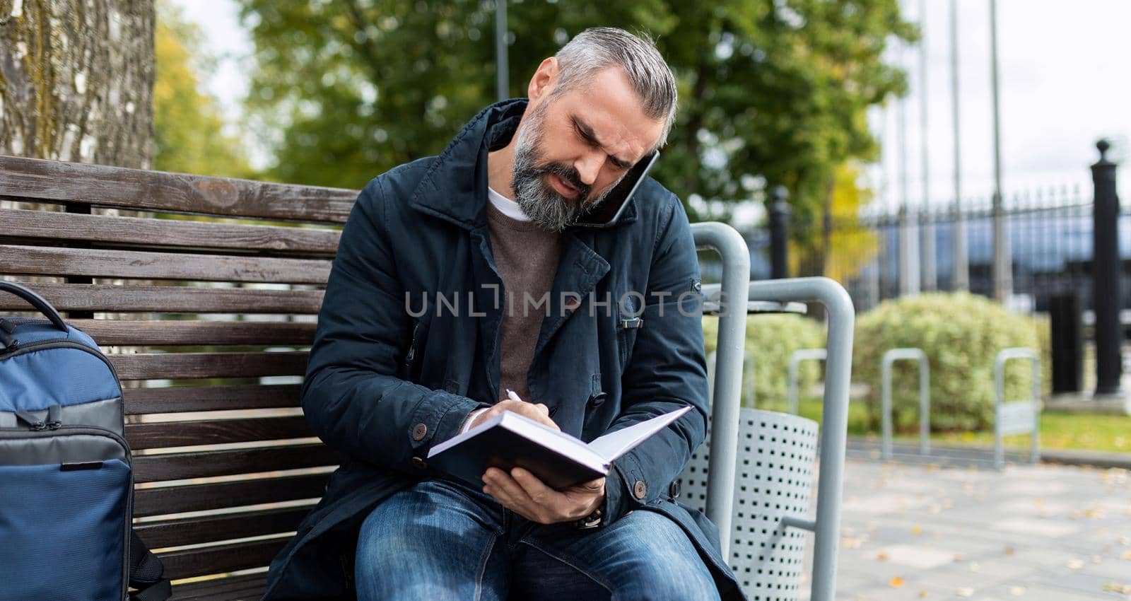 businesswoman talking on the phone making notes in a notebook while sitting on a bench in a city park.