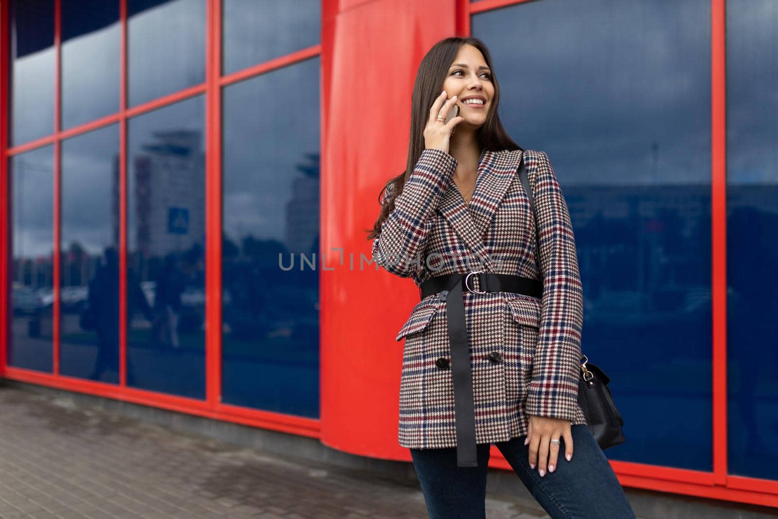 a young woman in an autumn coat speaks on a mobile phone against the background of a red blue building facade.