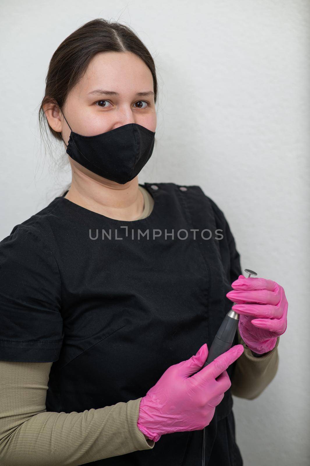 The pedicure master stands against the background of a white wall and holds an apparatus with an abrasive disc