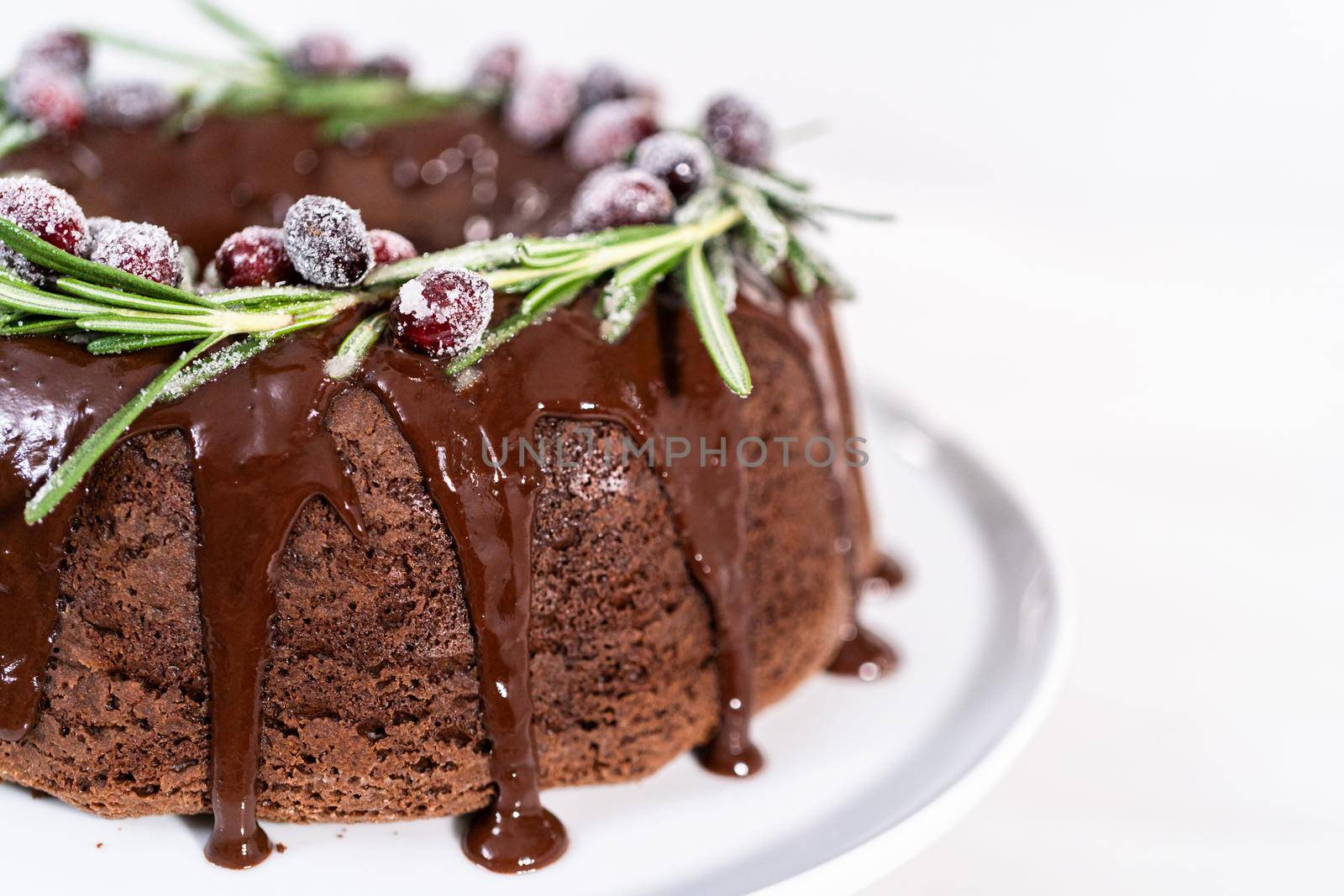 Chocolate bundt cake with chocolate frosting decorated with fresh cranberries and rosemary covered in a white sugar.