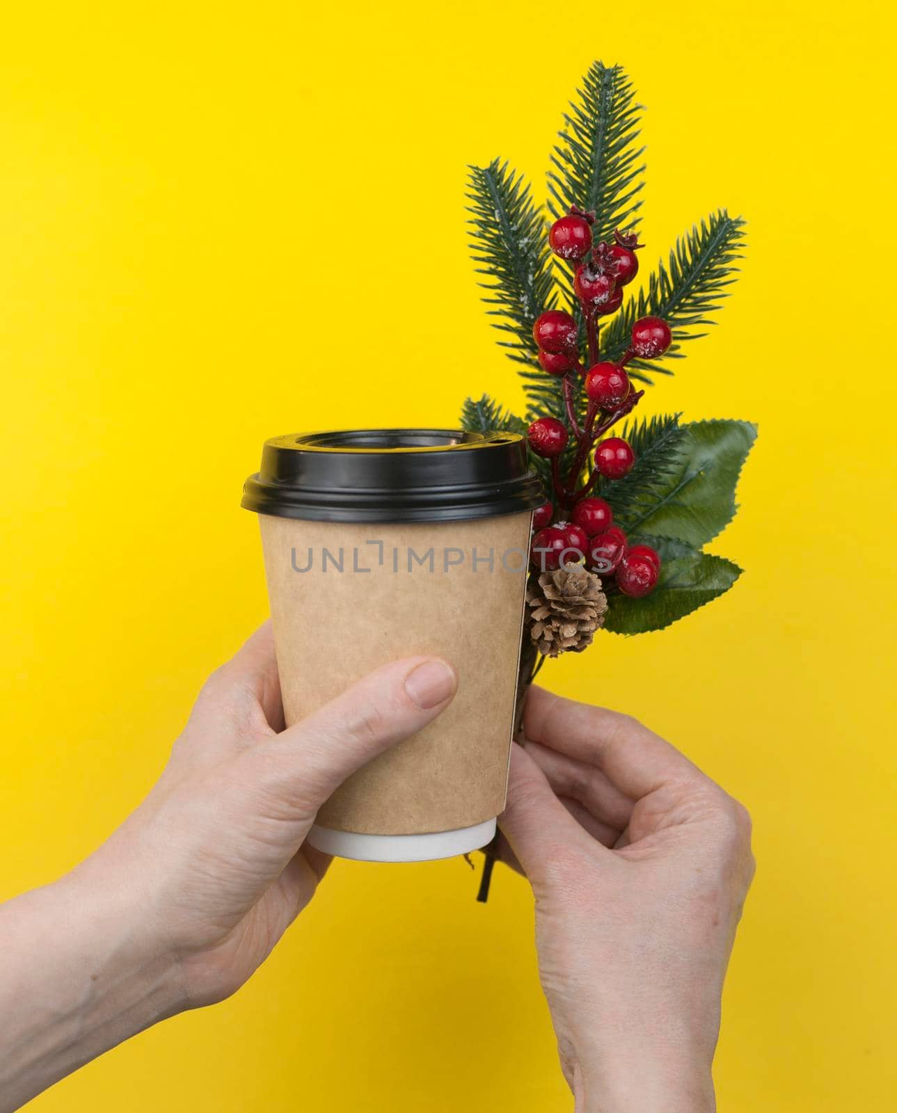 disposable coffee cup in woman hand with Christmas berry branches on yellow background. Concept Christmas. Copy space. 