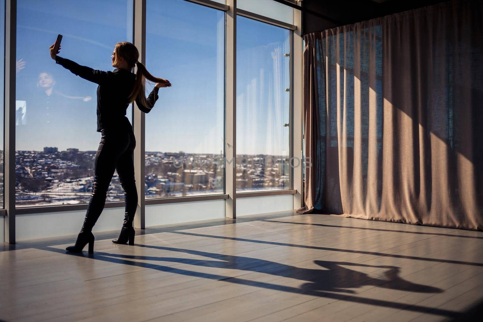 A beautiful girl takes a selfie on her phone against the background of panoramic windows in a skyscraper