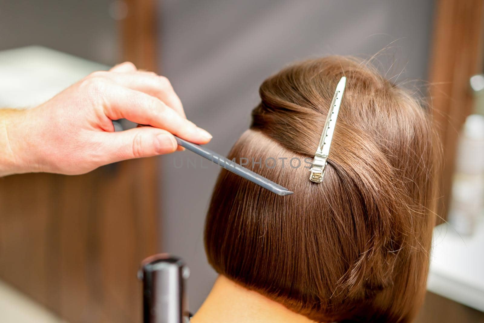 Hairstylist is straightening short hair of young brunette woman with a flat iron in a hairdresser salon, close up