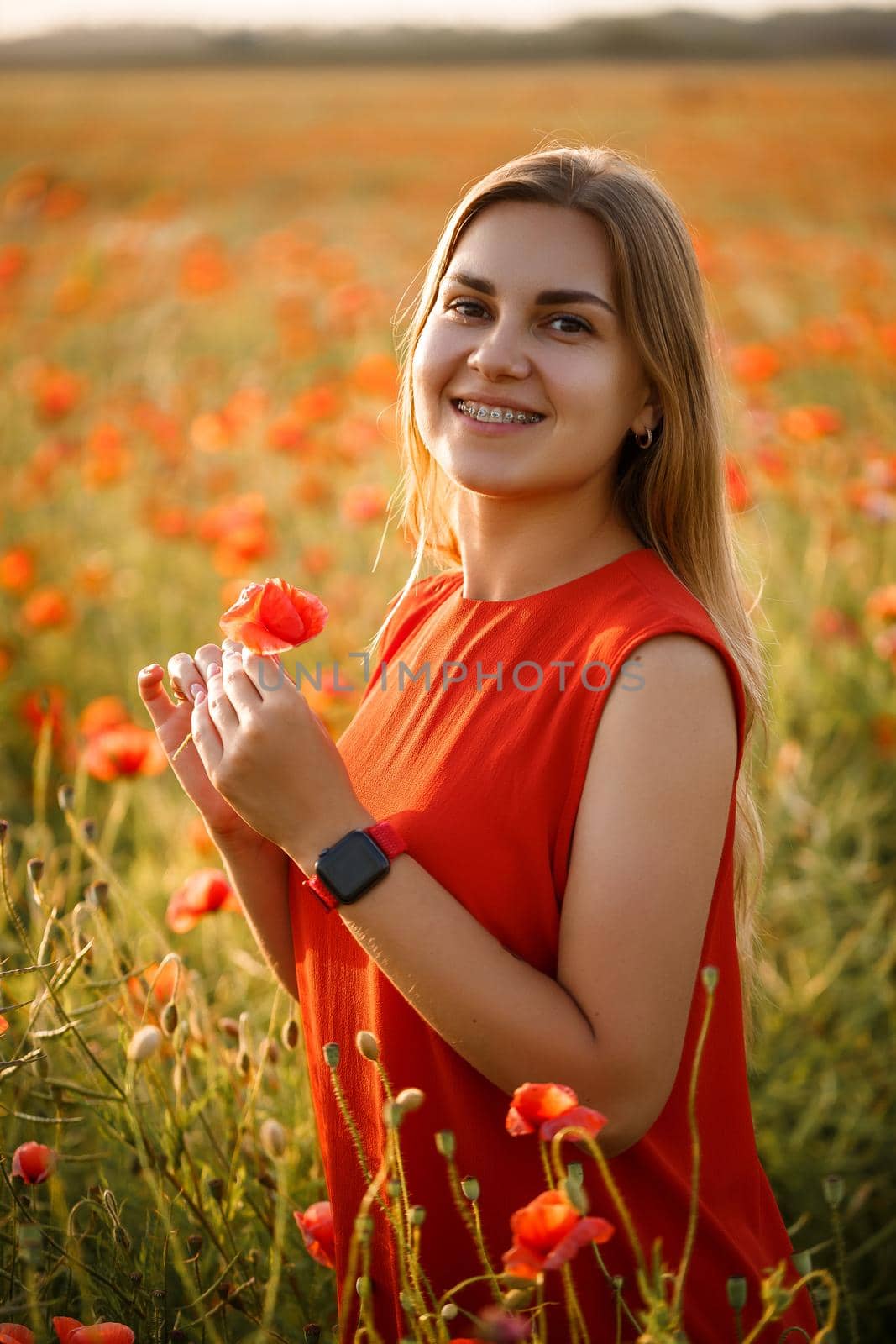 Happy blonde woman in red dress holding a flower in her hair on a poppy field. Fun summer vibes. Relaxed atmosphere, positive emotions.