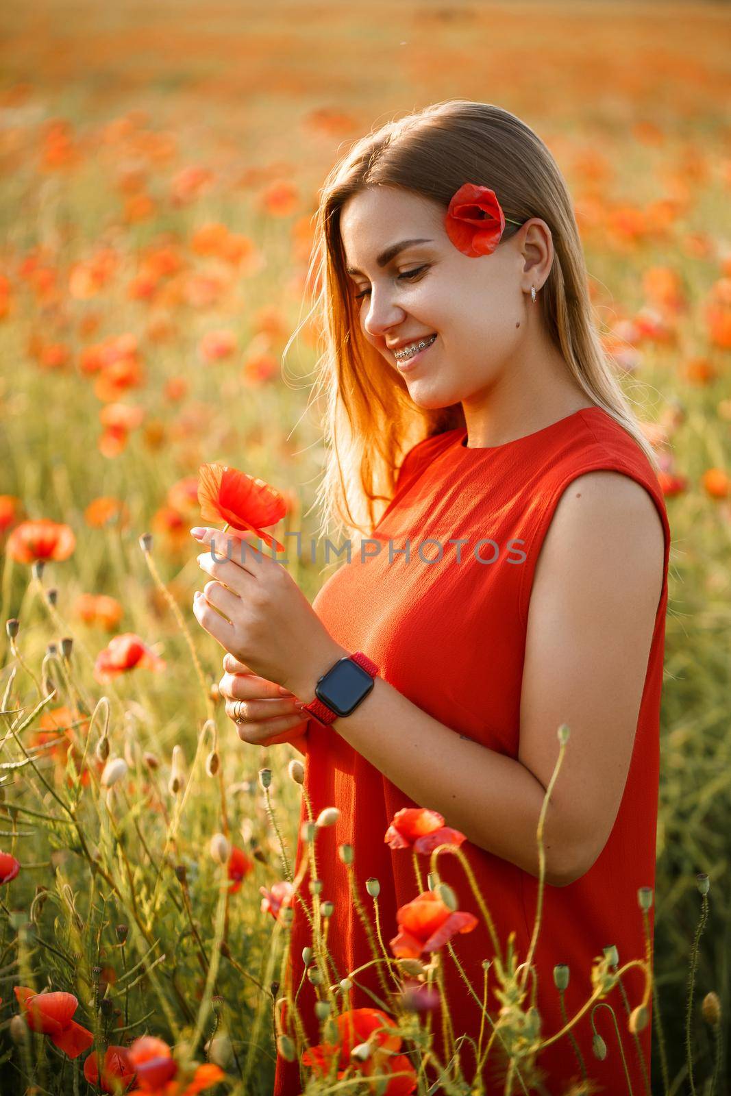 Happy blonde woman in red dress holding a flower in her hair on a poppy field. Fun summer vibes. Relaxed atmosphere, positive emotions.