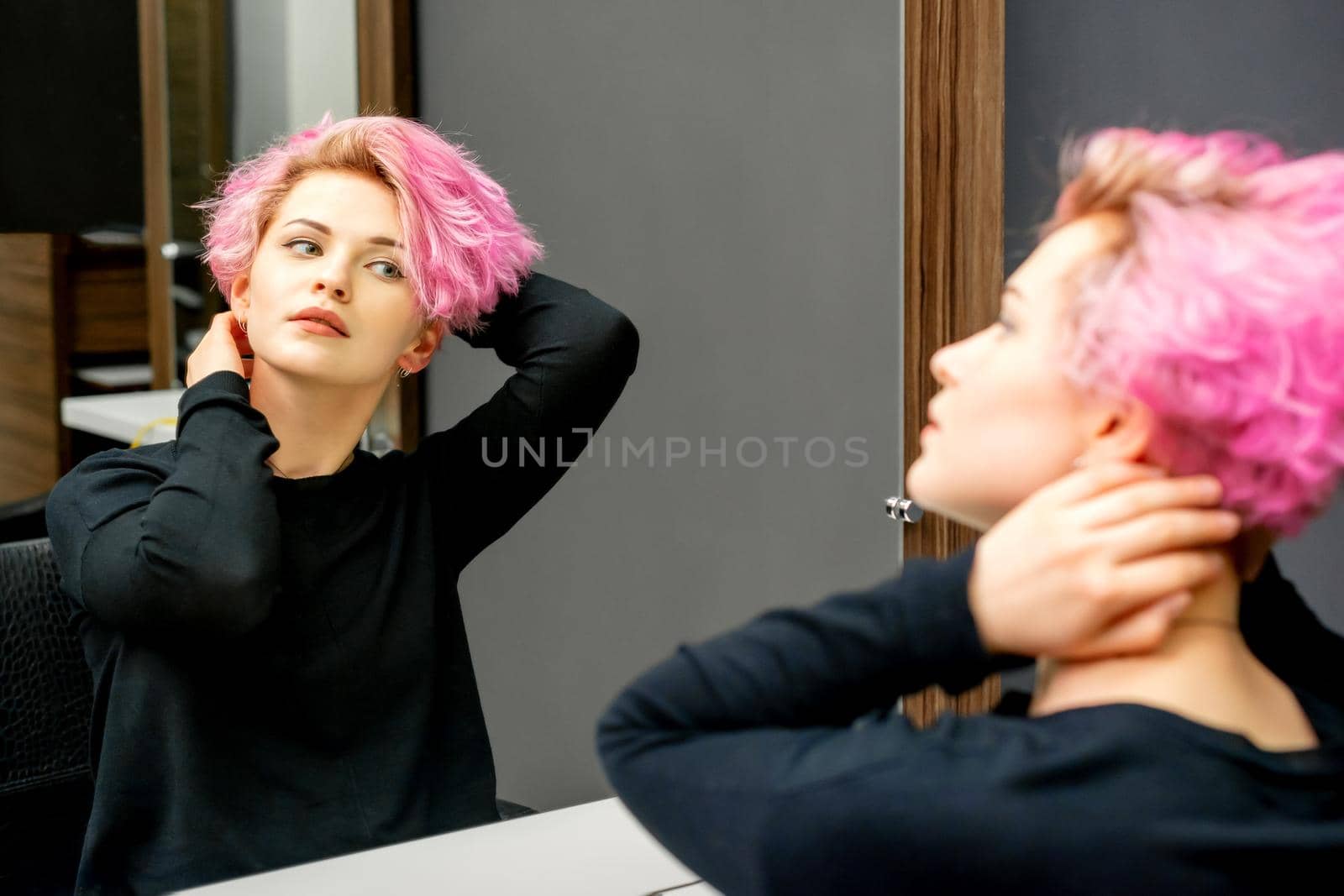 The beautiful young caucasian woman with a new short pink hairstyle looking at her reflection in the mirror checking hairstyle in a hairdresser salon