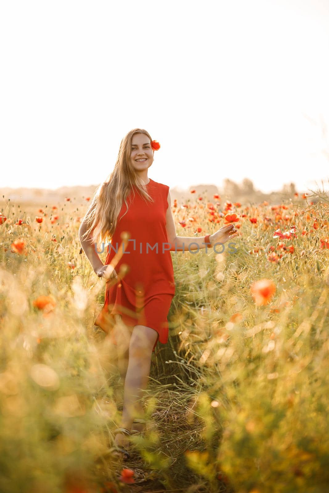 A woman of European appearance with long blonde hair and a red summer dress, she stands in a blooming poppy field