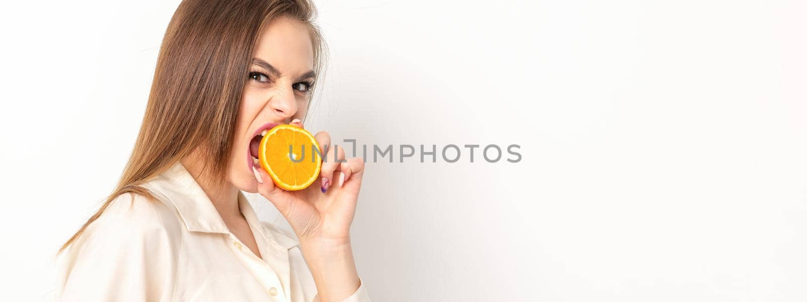 Young caucasian pretty cunning brunette woman biting one orange half and looking at the camera wearing a white shirt over white background