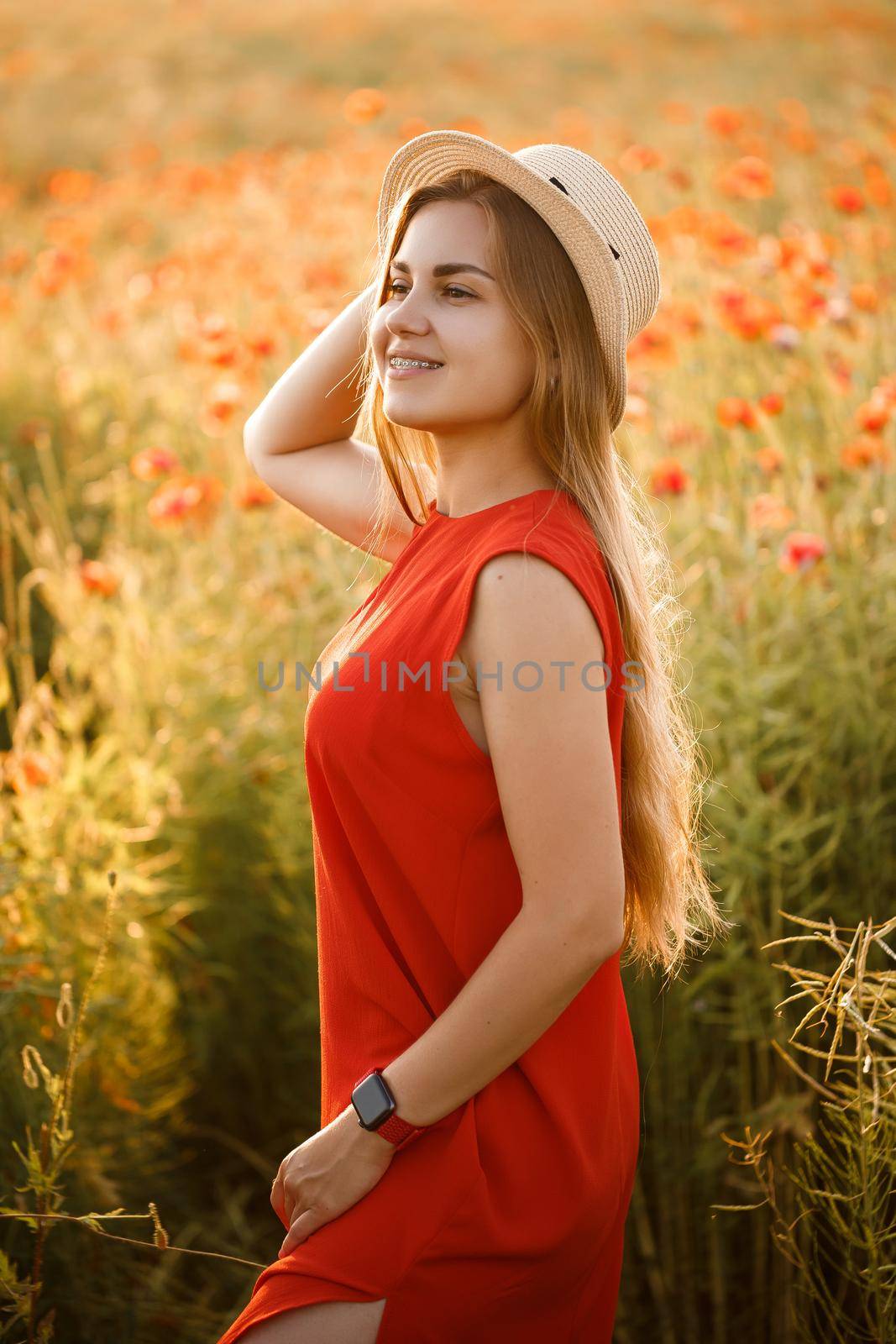 Happy cheerful woman walking on blooming field poppies. Red flowers. A blonde in a red short dress and a hat.