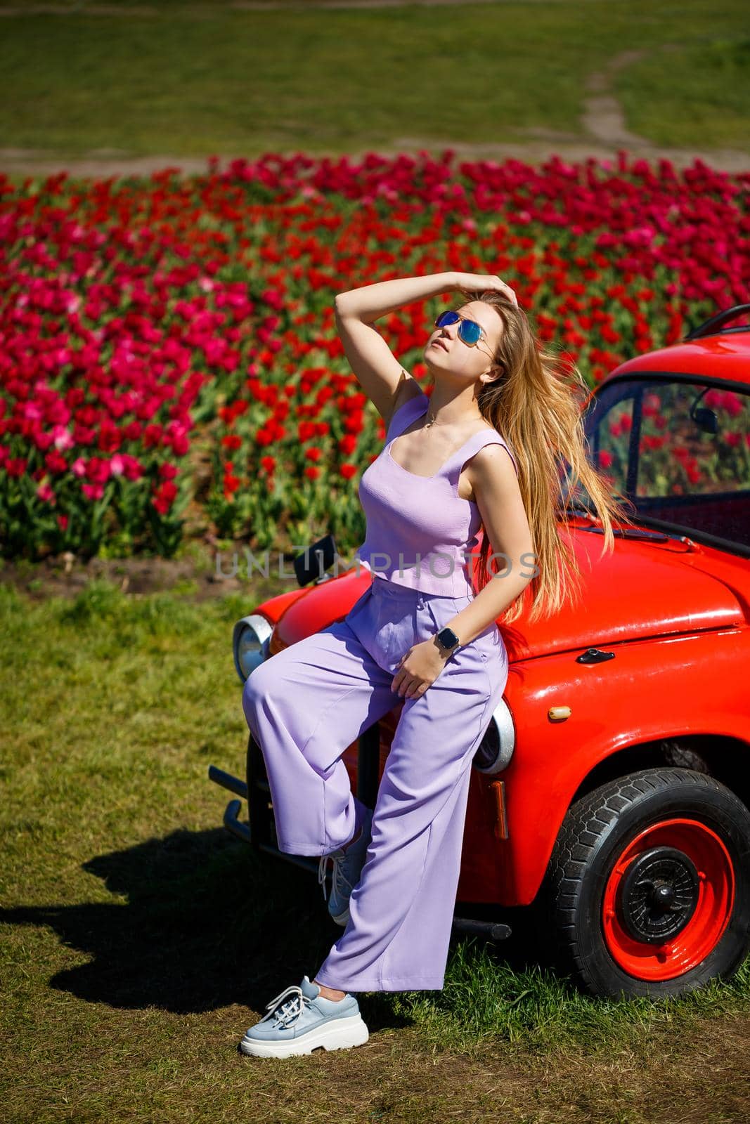 A girl model in casual clothes sits on a red car among tulip fields.