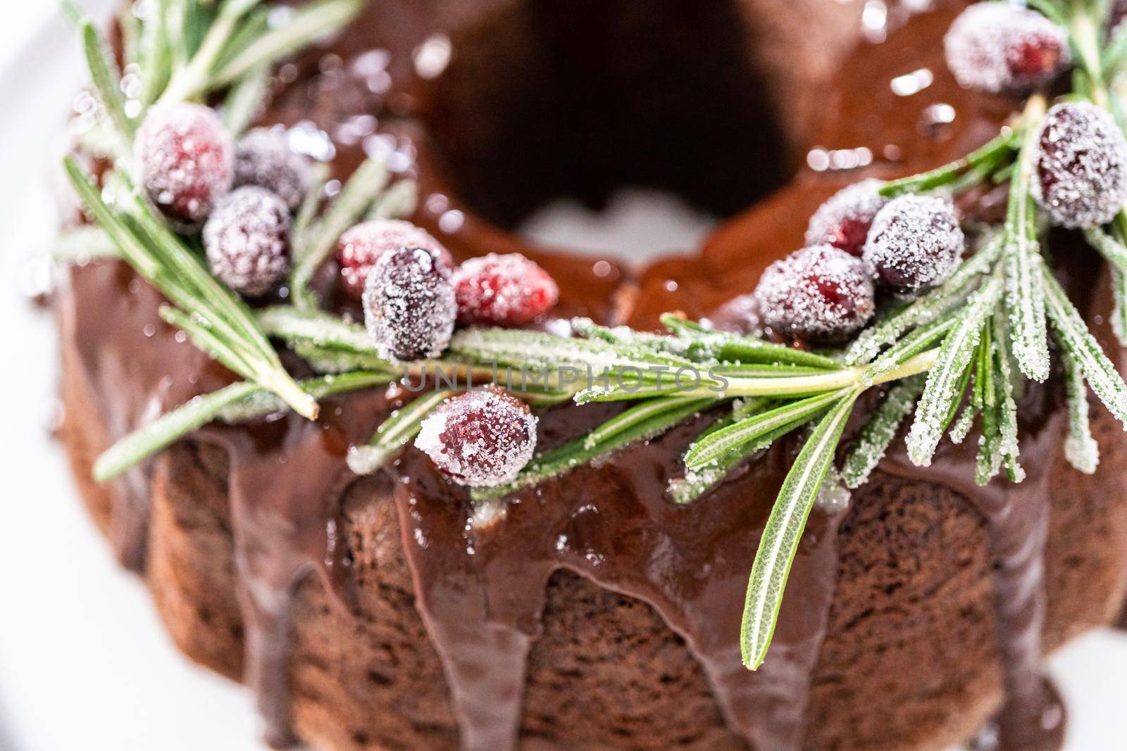 Chocolate bundt cake with chocolate frosting decorated with fresh cranberries and rosemary covered in a white sugar.