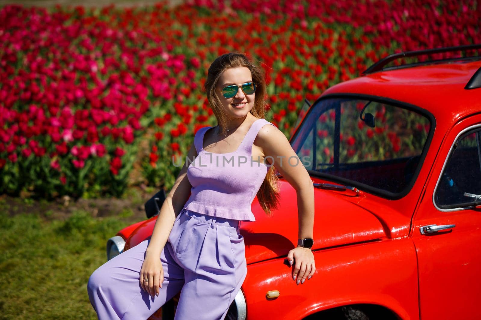 A girl model in casual clothes sits on a red car among tulip fields.