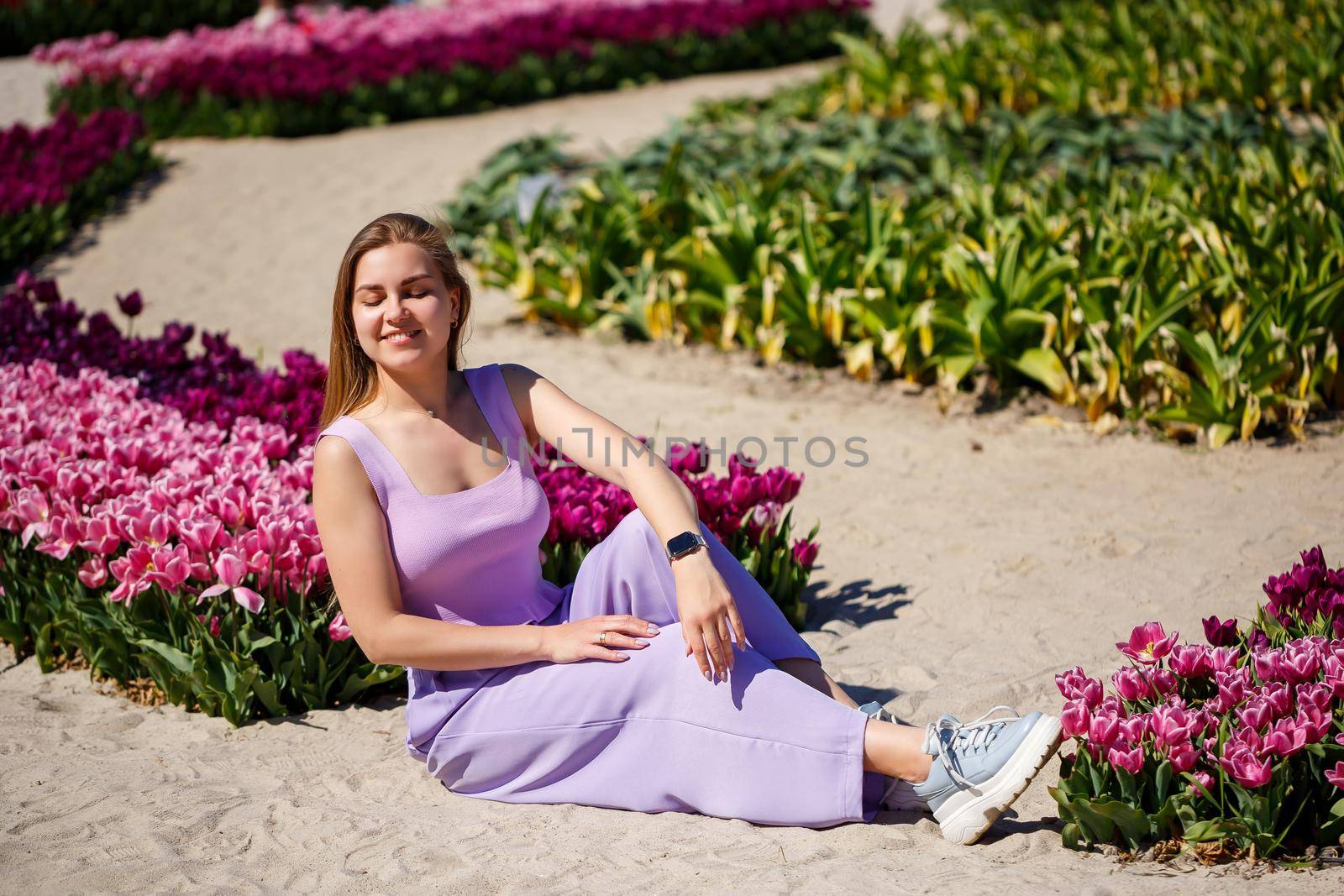 Back view of anonymous woman in summer dress and hat walking near blooming tulips in meadow on sunny day in nature