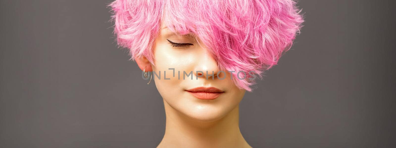 Beautiful young caucasian woman with short curly bob hairstyle dyed in pink color with closed eyes against dark gray background with copy space