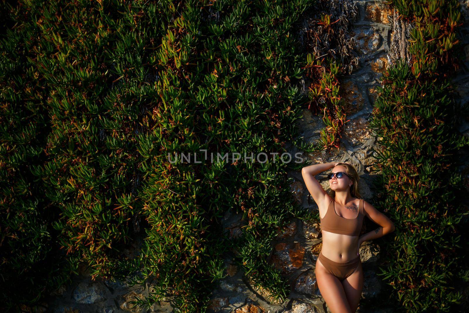 Beautiful sexy girl in sunglasses in a beige swimsuit. Young woman with tanned body soit near stone wall with green plants