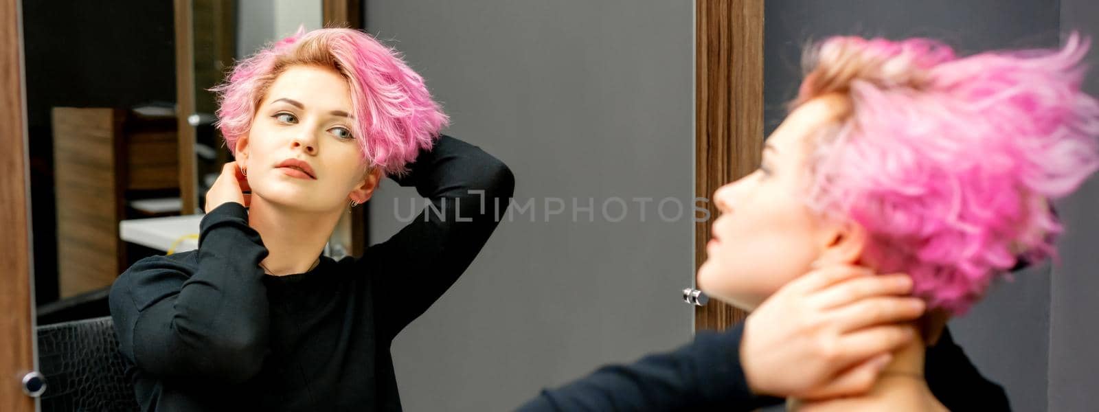 The beautiful young caucasian woman with a new short pink hairstyle looking at her reflection in the mirror checking hairstyle in a hairdresser salon