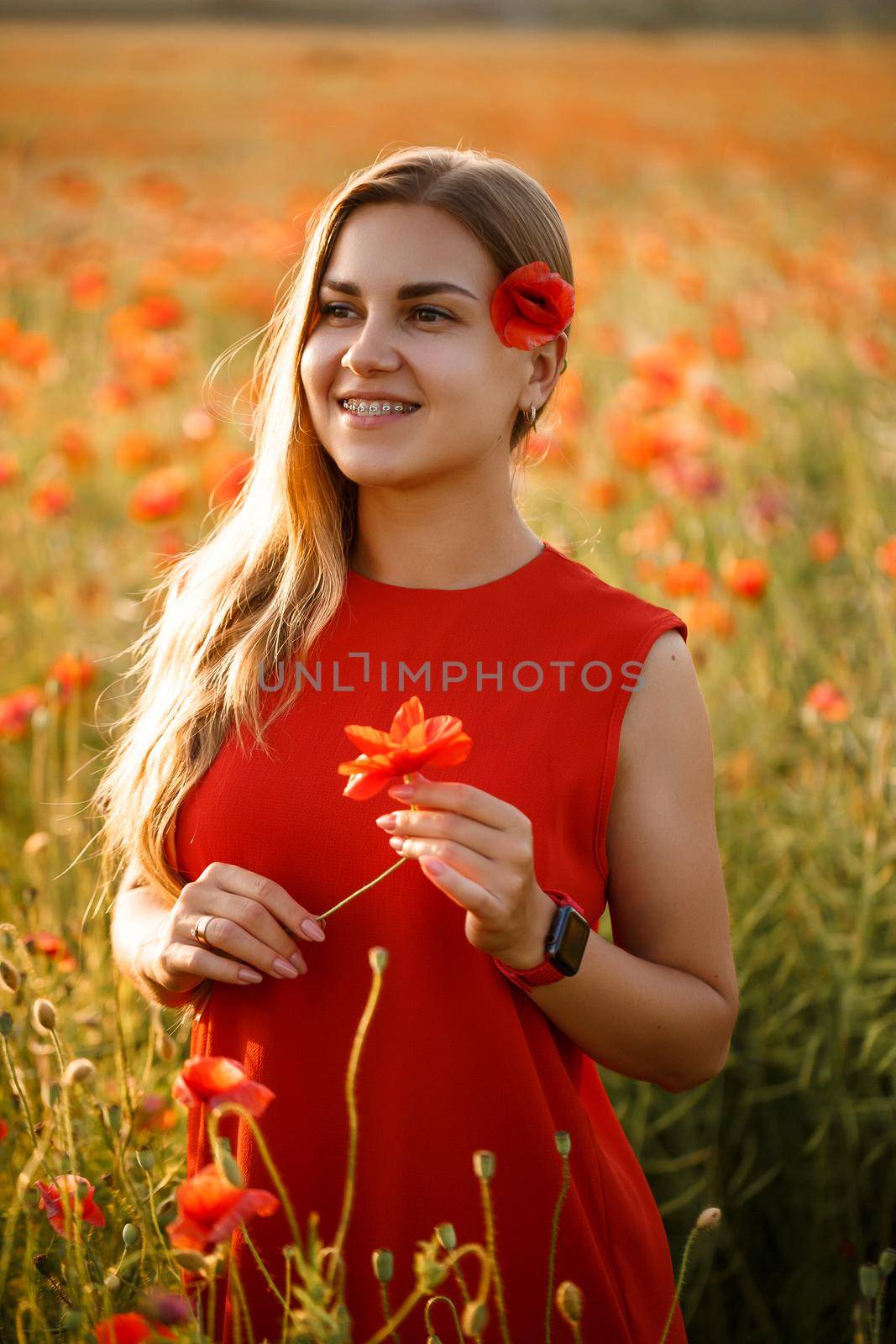 Happy blonde woman in red dress holding a flower in her hair on a poppy field. Fun summer vibes. Relaxed atmosphere, positive emotions.