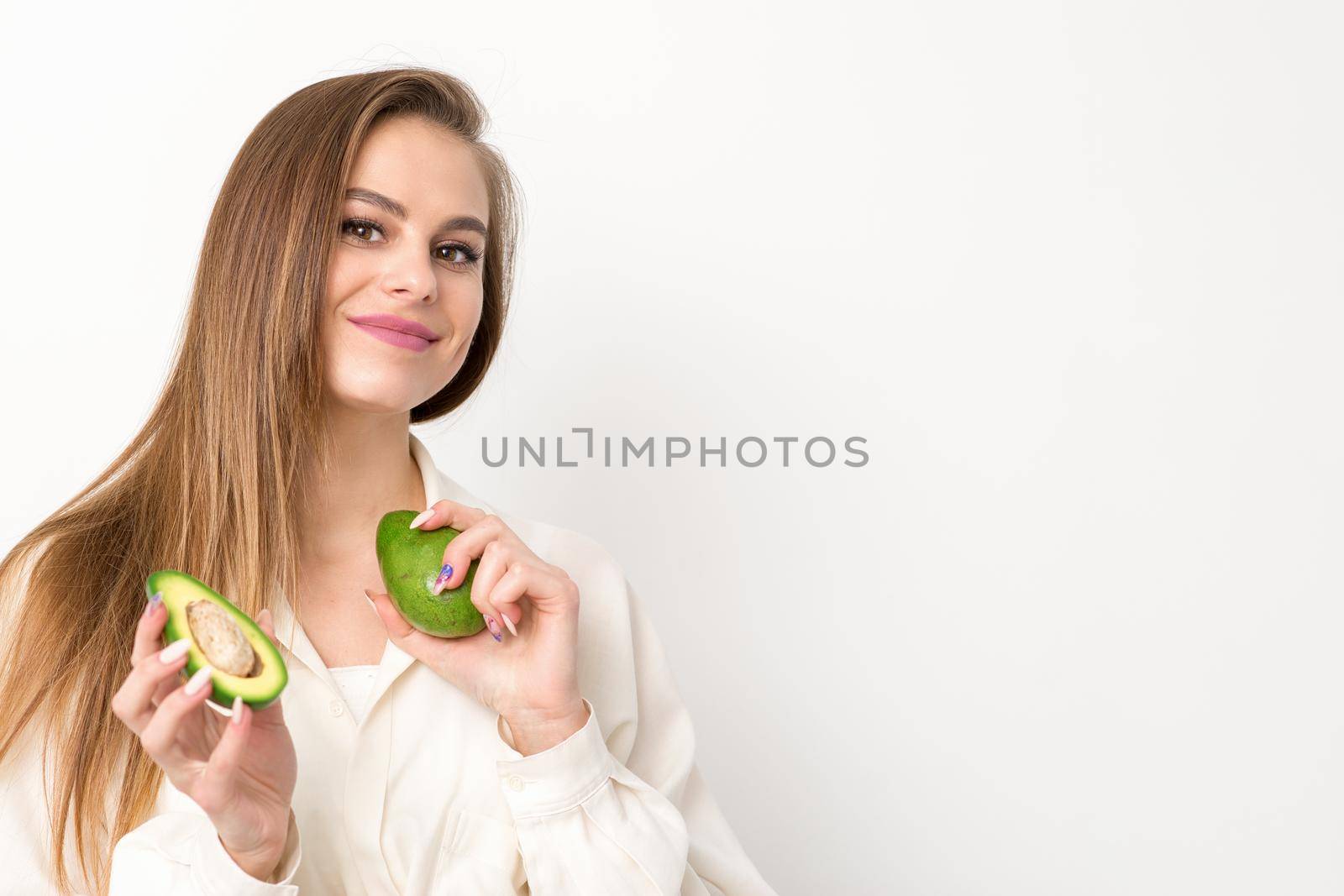 Portrait of a lovely smiling young brunette caucasian woman wearing the white shirt with long hair holding and showing avocado, standing isolated over white background