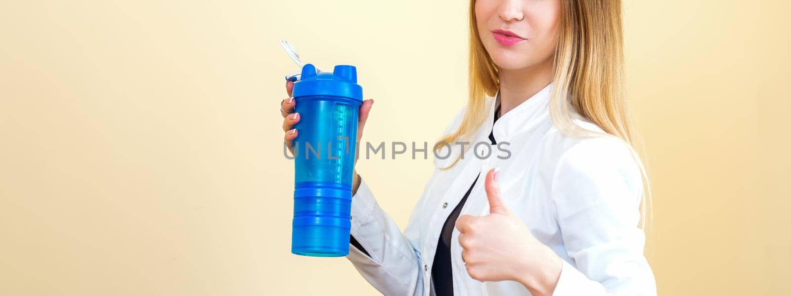 The young Caucasian female doctor wearing a white coat holds a blue plastic bottle with water with thumb up looking at the camera