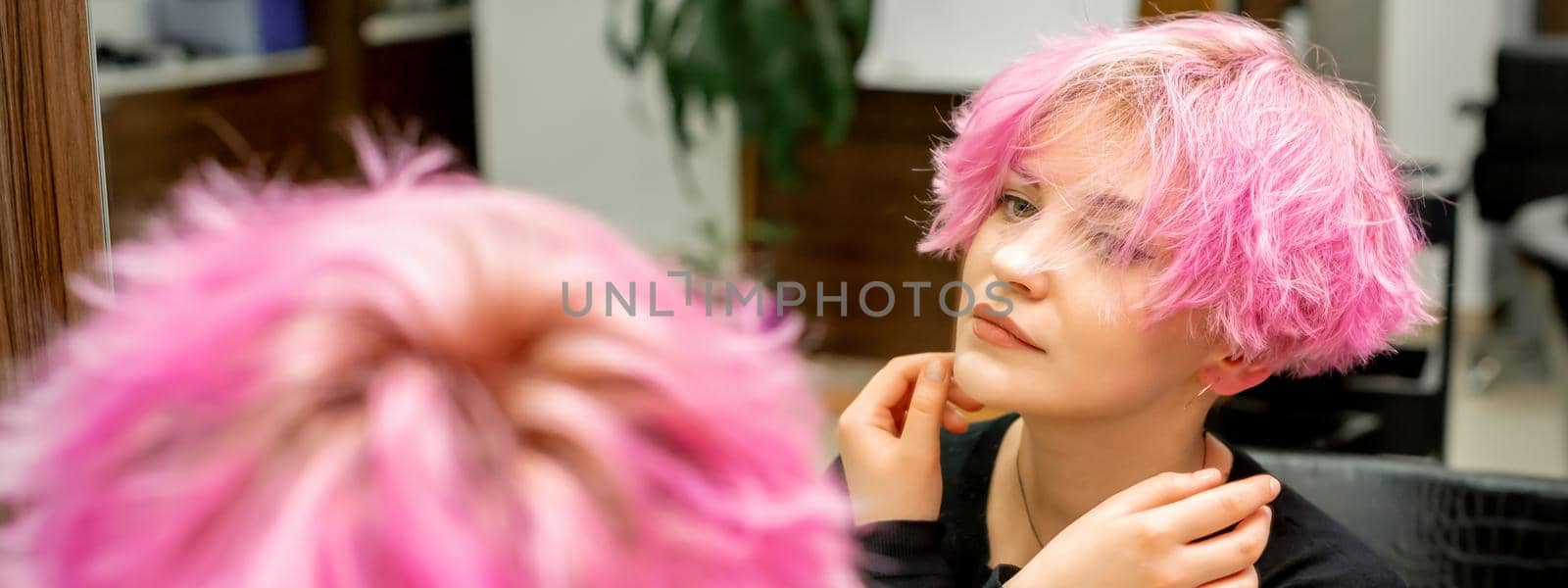 The beautiful young caucasian woman with a new short pink hairstyle looking at her reflection in the mirror checking hairstyle in a hairdresser salon