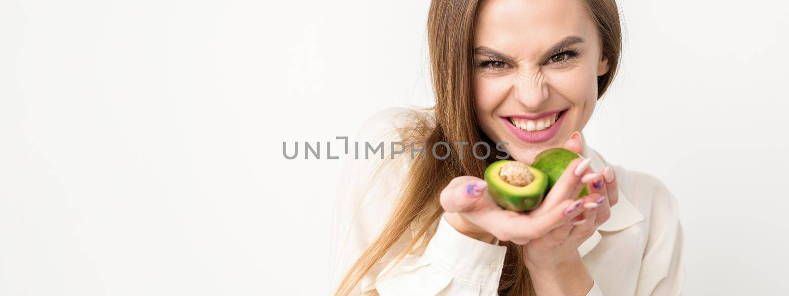 Portrait of a lovely smiling young brunette caucasian woman wearing the white shirt with long hair holding and showing avocado, standing isolated over white background