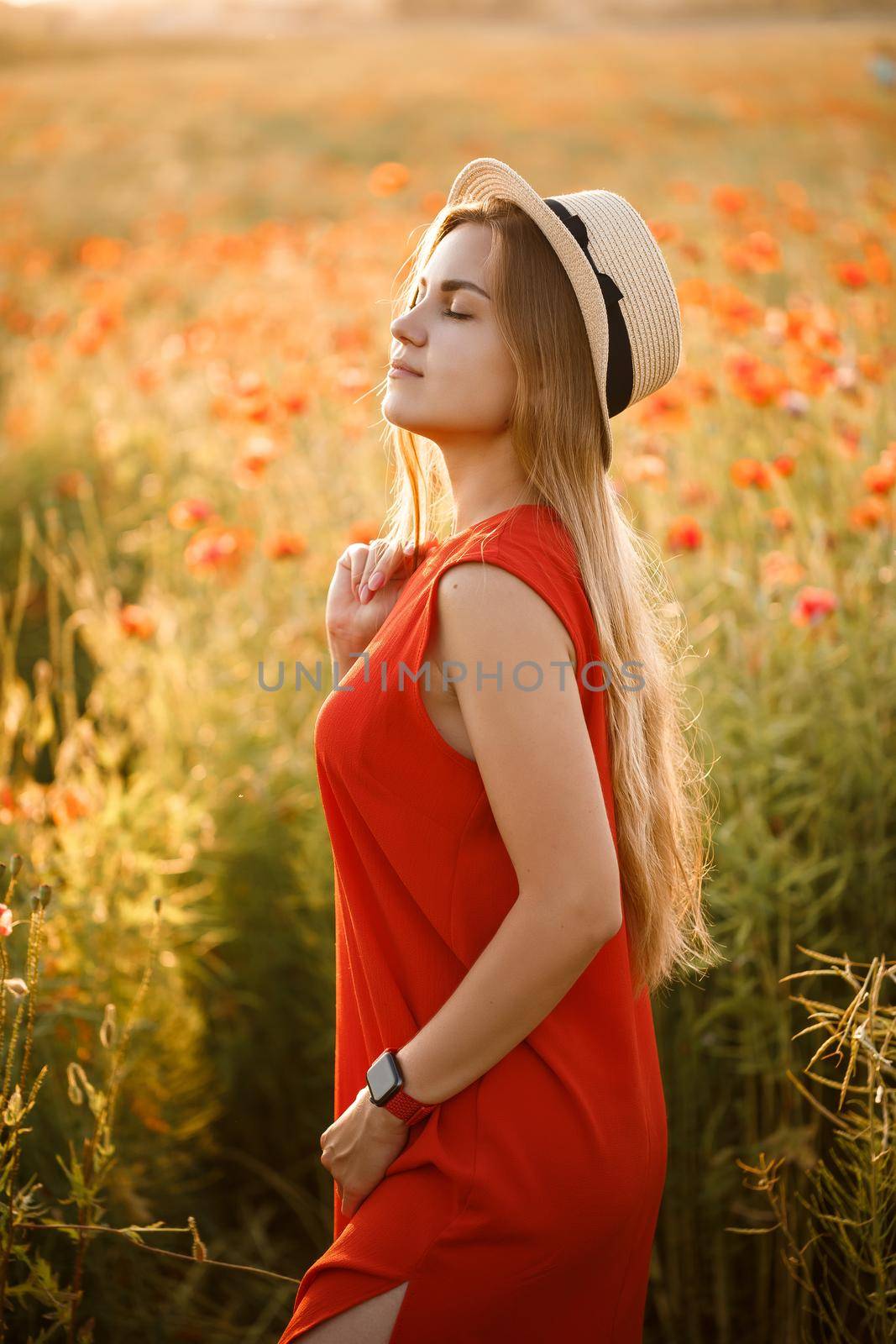 Happy cheerful woman walking on blooming field poppies. Red flowers. A blonde in a red short dress and a hat.