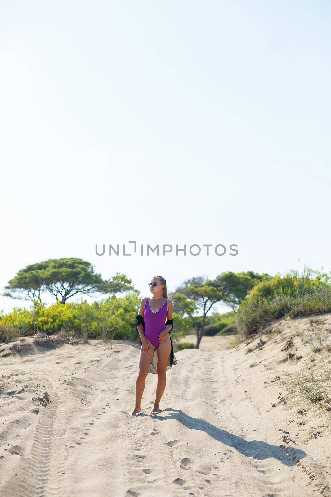 A slender tanned young woman in a swimsuit and a pareo, standing on the golden sand in the desert. Model woman resting and sunbathing on vacation. Travel concept, swimwear