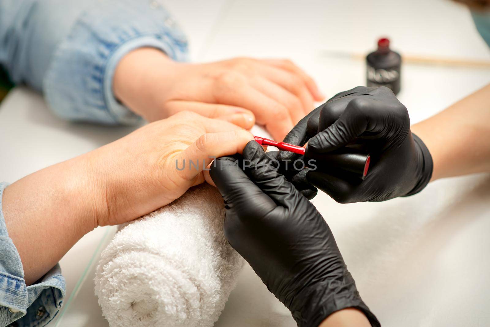 Manicure varnish painting. Close-up of a manicure master wearing rubber black gloves applying red varnish on a female fingernail in the beauty salon. by okskukuruza