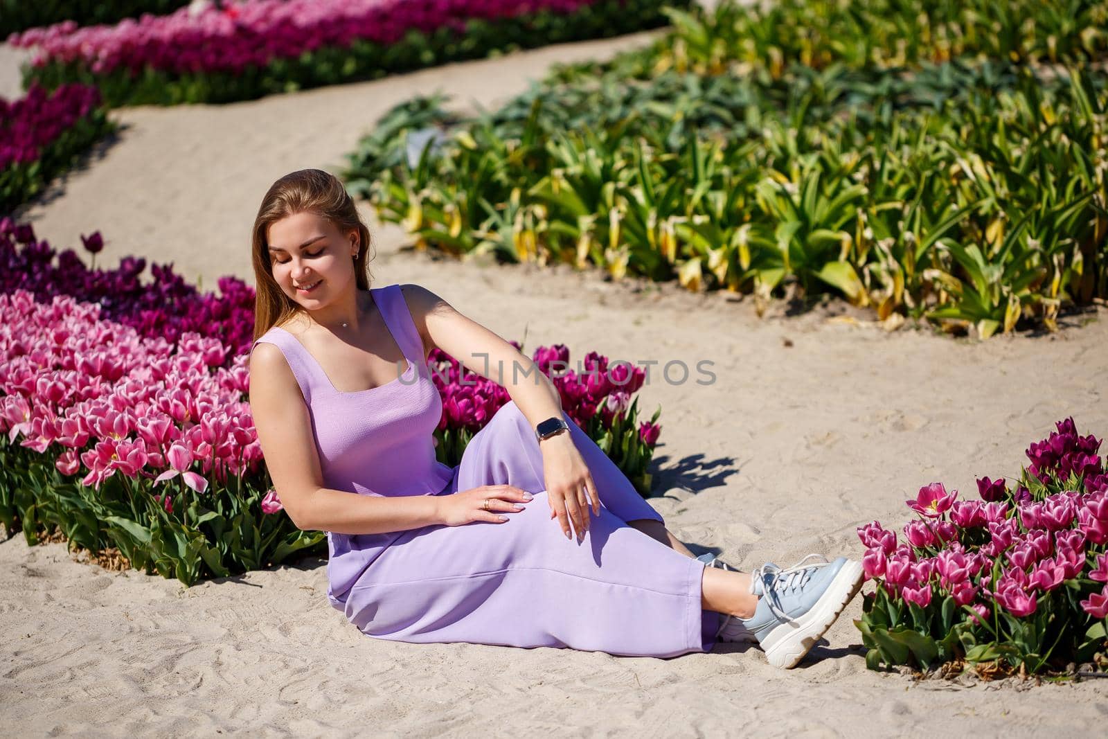 Back view of anonymous woman in summer dress and hat walking near blooming tulips in meadow on sunny day in nature