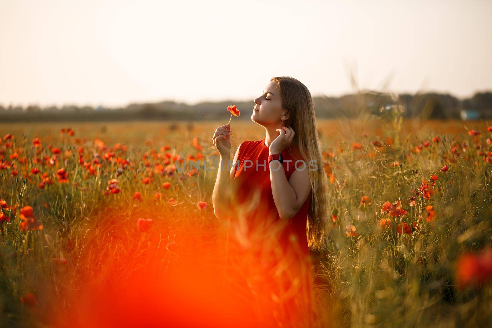 A woman of European appearance with long blonde hair and a red summer dress, she stands in a blooming poppy field