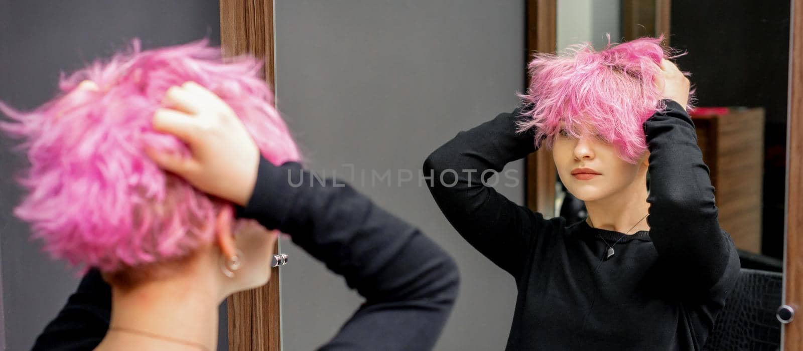 Young woman checking her new curly short pink hairstyle with he hands in front of the mirror at the hairdresser salon.