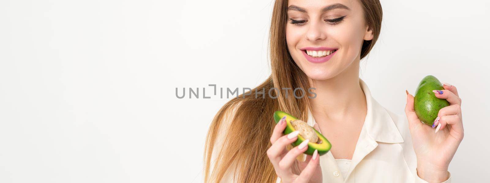 Portrait of a lovely smiling young brunette caucasian woman wearing the white shirt with long hair holding and showing avocado, standing isolated over white background